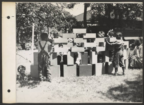 Image: A black and white outdoor shot on March 20, 1936, of several preschool children playing with and constructing a large structure made of large rectangular blocks. The children interact; some are standing, and others are working with the blocks, while others are sitting in the grass among the pieces. An adult is swatting near the children on the far right corner. At least one child is wearing overalls with a shoulder strap design on their top. Trees are visible in the background, and a portion of a building is seen behind the children. Photo Credit: United States Works Progress Administration, Courtesy of Chicago Park District Records: Photographs at the Chicago Public Library. Special Collections & Preservation Division, Digital Link.