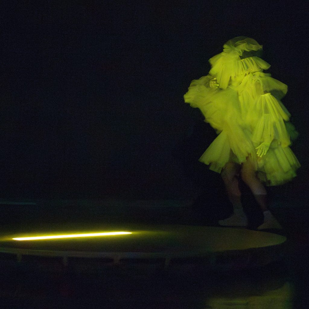 Image: Freeman | Po'Chop dances around a circular platform on the green-yellow lit stage wearing a fluffy dress and white gloves. Photo by Natasha Moustache.
