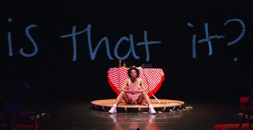Image: Freeman | Po'Chop sits on a wood platform on the stage. The wall says "is that it?". Photo by William Frederking.
