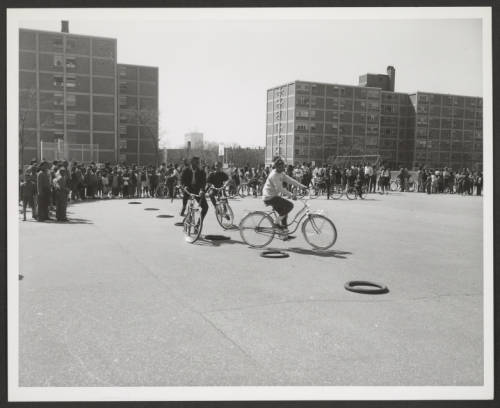 Image: A black and white photograph taken in 1965 of three young African Americans in the public housing courtyard of Madden Park Homes. Two large project buildings and a large and dense crowd surround them in the backdrop. Going in a large circle, one cyclist is slightly ahead of the other two. The cyclists appear to be Black men and appear to be young adults. Two large, multi-story brick apartment buildings are visible in the background, indicating a densely populated urban environment. Photo Credit: unknown, Courtesy of Chicago Park District Records: Photographs at the Chicago Public Library. Special Collections & Preservation Division, Digital Link.