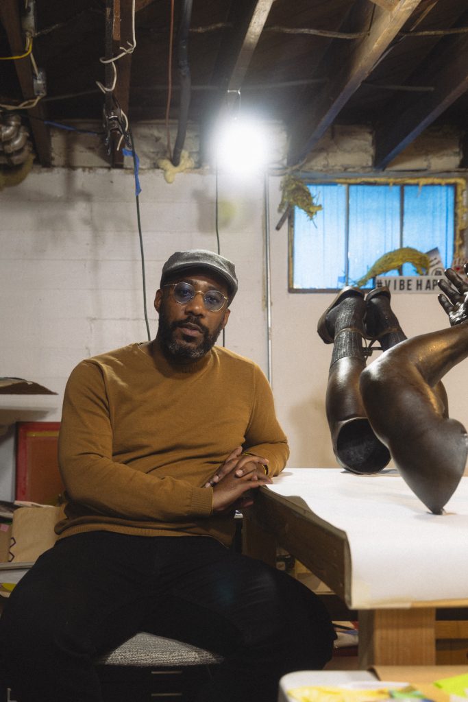Image: A portrait of Michael Coppage in his home studio, sitting on a chair next to his sculpture, Hands Behind Your Back, which depicts a hyper-realistic pair of arms and legs detained by zip ties.