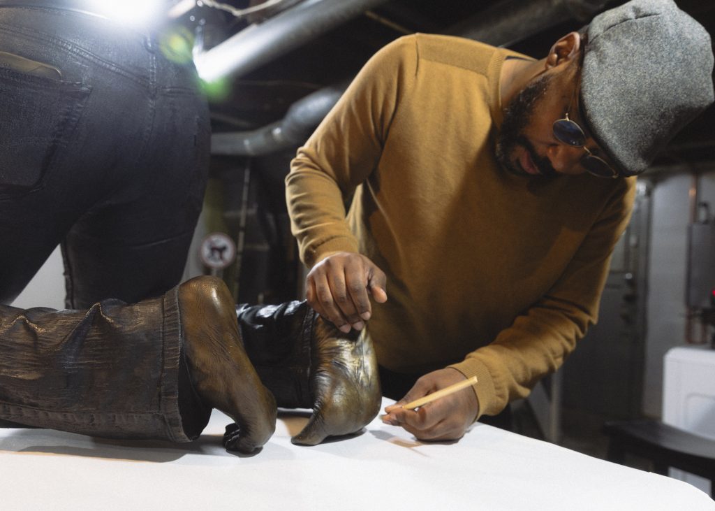 Image: Detail of Michael Coppage in his home studio handling his bronze sculpture, "Hands behind your back," depicting a hyper-realistic human waist and legs in a kneeling position. Michael stands at the back of the sculpture dusting the bare feet of the sculpture. Photo by Asa Featherstone, IV.