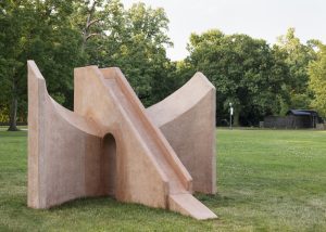 Image: Asad Raza. Orientation, 2022. Photography by Field Studio. Courtesy of the artist. A photo of a large sculptural piece outside in the grass. The piece is monochromatic (a light terracotta color) and is geometric in shape.