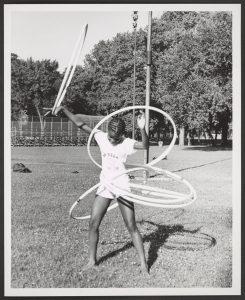 Image: A black and white photograph of a young African American girl skillfully playing with five hula hoops simultaneously on July 15, 1974. She stands in the grass at McKinley Park, with mature trees, the gate of a baseball field, and a tall pole in her background. She wears a short-sleeved shirt and shorts. Her hair is styled with pattern braids. Photo Credit: unknown, Courtesy of Chicago Park District Records: Photographs at the Chicago Public Library, Special Collections & Preservation Division.