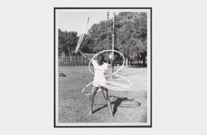 Image: A black and white photograph of a young African American girl skillfully playing with five hula hoops simultaneously on July 15, 1974. She stands in the grass at McKinley Park, with mature trees, the gate of a baseball field, and a tall pole in her background. She wears a short-sleeved shirt and shorts. Her hair is styled with pattern braids. Photo Credit: unknown, Courtesy of Chicago Park District Records: Photographs at the Chicago Public Library. Special Collections & Preservation Division, Digital Link.