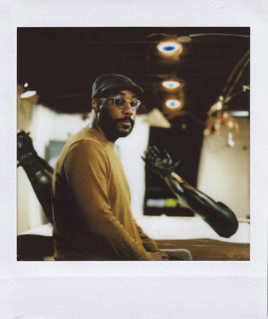 Image: Polaroid image of Michael Coppage sitting next to a bronze sculpture depicting a pair of hyper-realistic disembodied hands and legs tied by zip-ties. Photo by Asa Featherstone, IV.