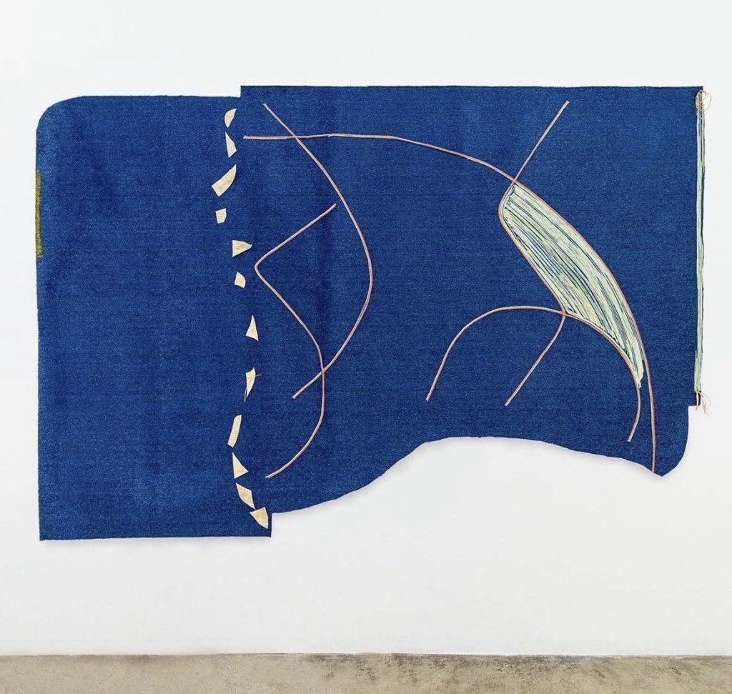 Image: Teresa Baker. Missouri River, 2022. Photography by Jacob Philip. Courtesy the Artist & de boer, Los Angeles & Antwerp. A large, amorphic piece hangs in a gallery on a white wall. The piece is largely deep blue with various lines and abstract shapes, all in earth tones.