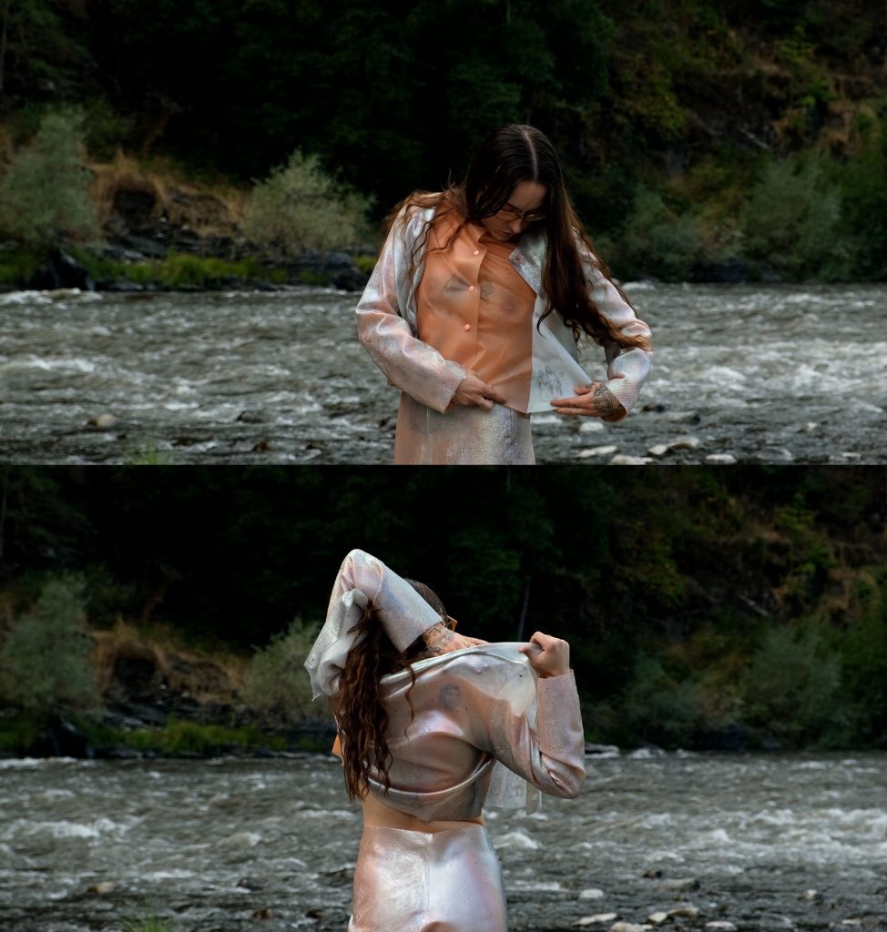 In the upper still, Robinson is standing in the river facing the camera and reaching across their body to adjust the jacket and tan silicone undershirt. In the bottom still, Robinson is standing in the river with their back to the camera and is reaching back with their left arm to adjust the jacket under their right shoulder. 