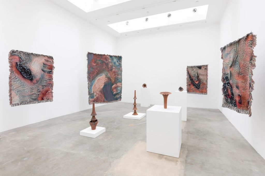 Four AI-generated textiles hang in a white gallery space with several sculptures displayed on plinths in the center of the room. 