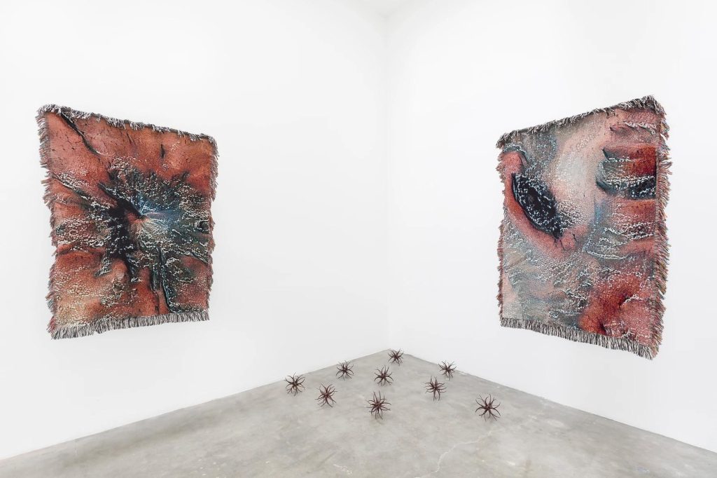 Two AI-generated textiles hang on opposite walls with nearly a dozen small sculptures displayed on the floor. 