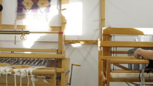 Image: A photograph of two large textile looms side by side in a bright, sun-lit room. A person's arm can be seen working with materials on one loom, while the other loom displays a piece in progress. Photograph by Ian Carstens.