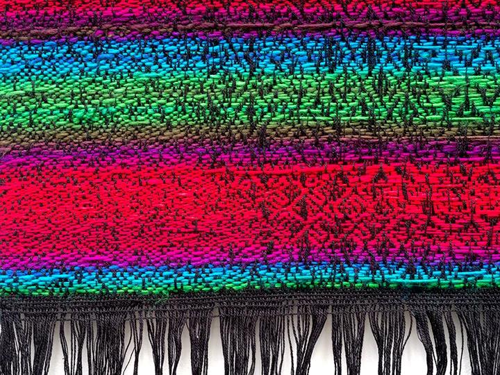 A close-up image of the edge of a pixellated, jacquard textile. The textile alternates between red, purple, blue, green with a black edge. 
