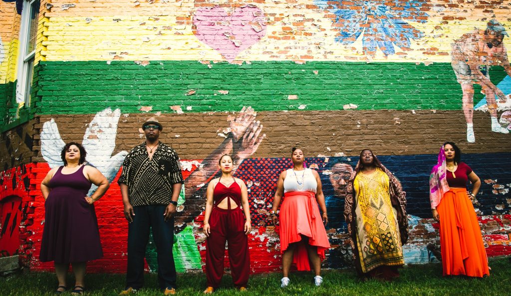 Image: A photograph of six members of Honey Pot Performance. They all stand in a row facing the camera in front of a mural on a brick wall. Photo by Marcus Davis, courtesy of Honey Pot Performance.