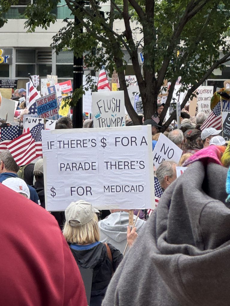 Image: A central sign, in the midst of a crowd, reads: "If there's $ for a parade, there's $ for Medicaid." The first dollar sign is upside-down. Image courtesy of the author.