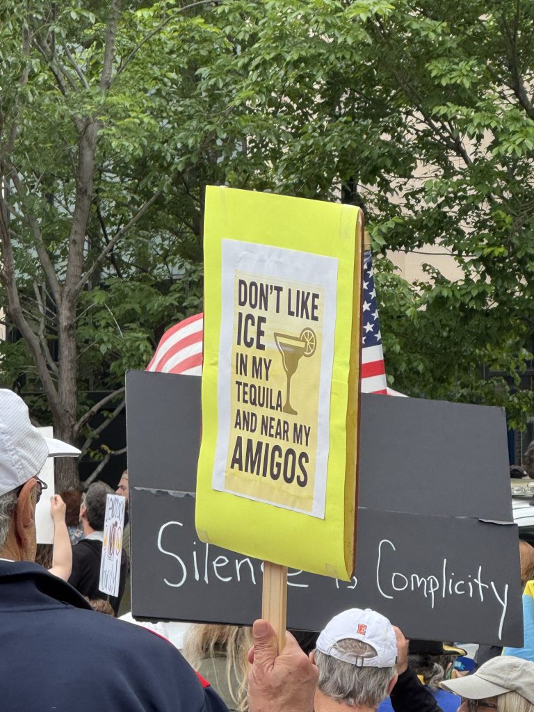 Image: A yellow sign and a black sign are held up perpendicular to each other, The yellow one says "Don't like ice in my tequila and near my amigos," and the black one says, "Silence is complicity." Image courtesy of the author.