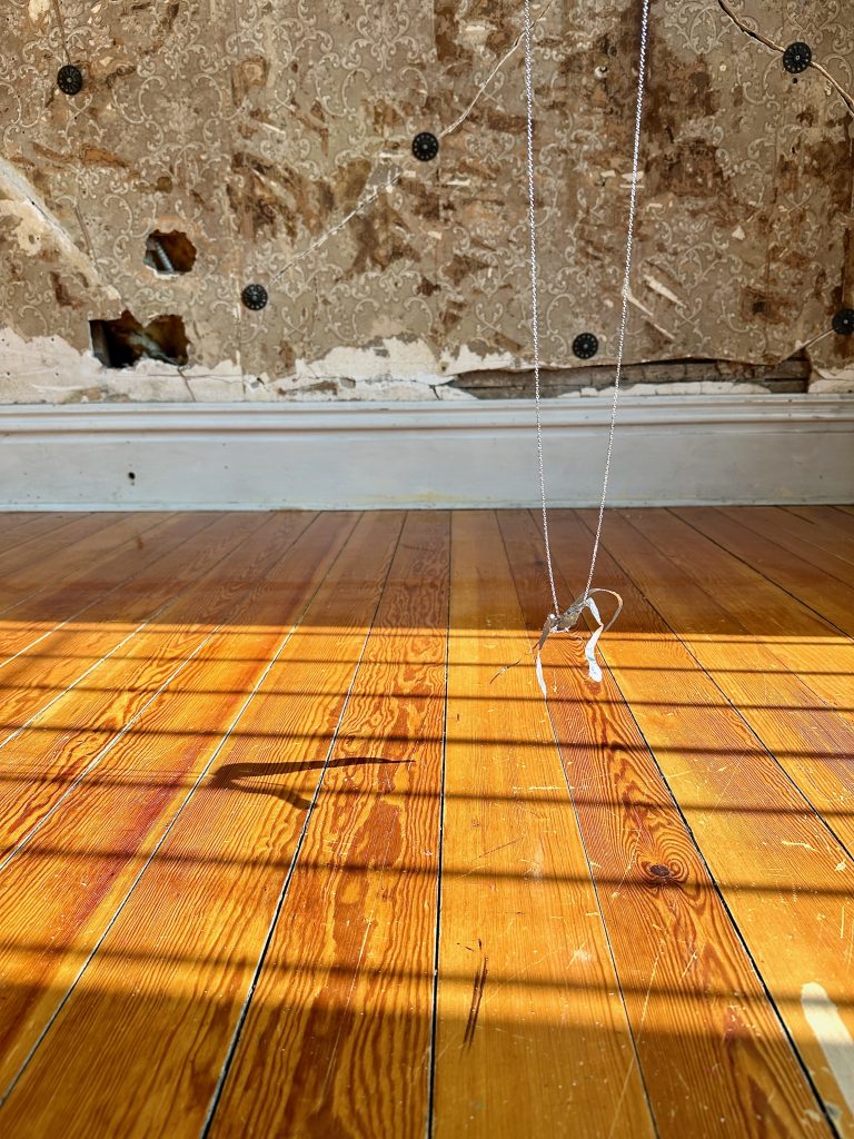 Image: Blas Isasi, El viento will bring us home (detail), 2024. SHED Projects, Cleveland, OH. Details from Blas Isasi’s installation El viento will bring us home at SHED Projects. A small, delicate sculpture made from twisted metal and wire hovers just above the glowing wooden floor, casting a shadow in the gridded light of a window. Behind it, a partially peeled wall reveals floral wallpaper remnants. Photo courtesy of SHED Projects.