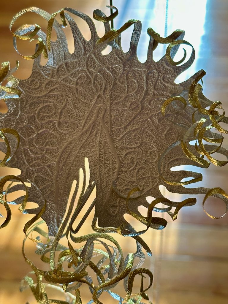 Image: Blas Isasi, El viento will bring us home (detail), 2024. SHED Projects, Cleveland, OH. Close-up detail of a suspended sculptural element from Blas Isasi’s El viento will bring us home. A finely cut, circular paper-like form is embossed with branching, organic patterns. The edges curl outward in metallic silver and gold tendrils, evoking vegetal or marine textures. The intricate form hangs delicately in space, partially backlit by window light. Photo courtesy of SHED Projects.