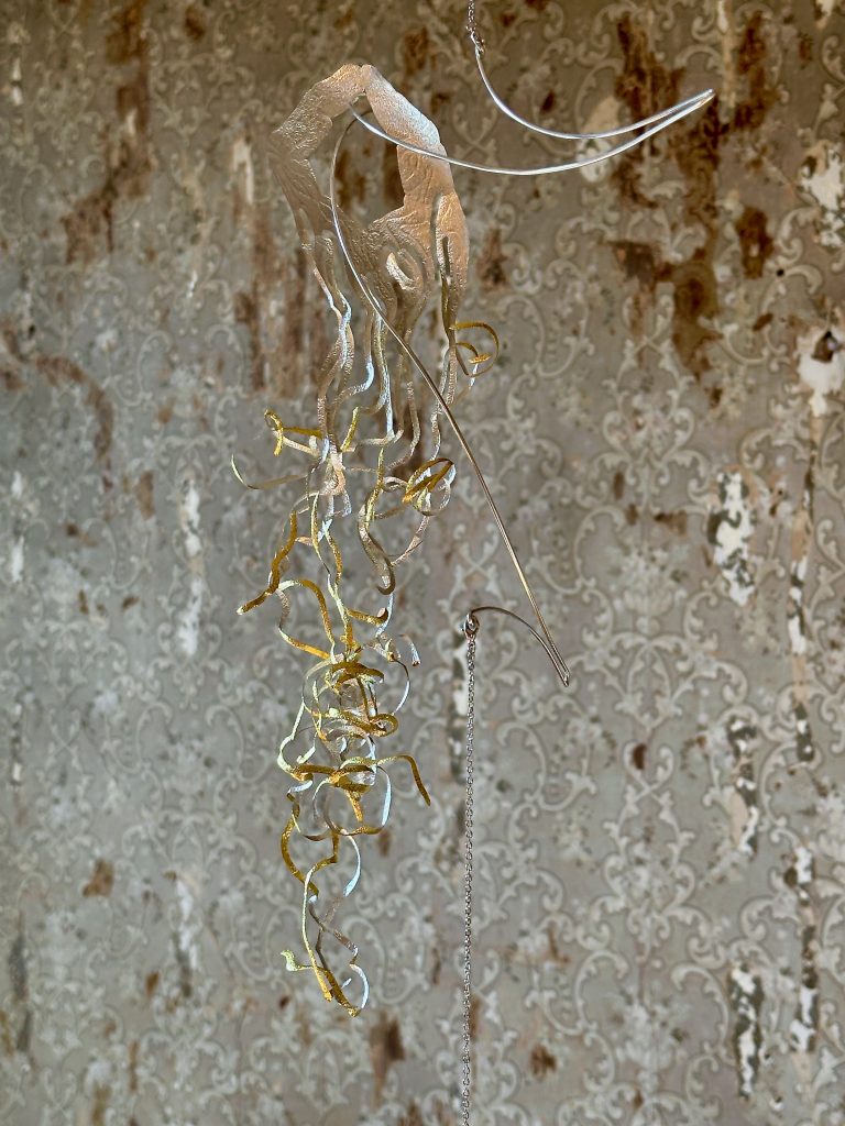 Image: Blas Isasi, El viento will bring us home (detail), 2024. SHED Projects, Cleveland, OH. Close-up of a suspended sculpture by Blas Isasi, part of El viento will bring us home at SHED Projects. A delicate form resembling a jellyfish or root system floats against a distressed wall with faded damask wallpaper. The top is composed of thin, translucent material, from which spiral tendrils of metallic gold cascade downward, interwoven with wire and clear rings. The background reveals peeling layers of history, emphasizing the installation's dialogue with architectural decay. Photo courtesy of SHED Projects.