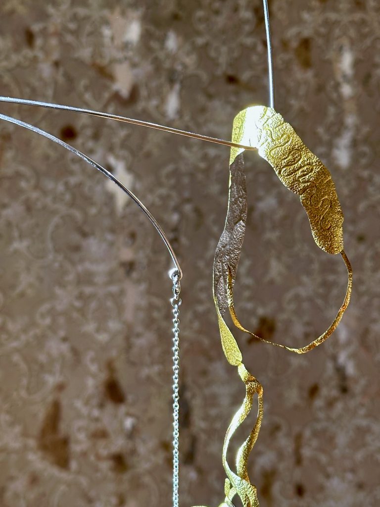 Image: Blas Isasi, El viento will bring us home (detail), 2024. SHED Projects, Cleveland, OH. Extreme close-up of a sculptural element from Blas Isasi’s El viento will bring us home. A thin, curling strip of textured gold foil in the shape of a finger is suspended alongside delicate wire and chain. The surface catches warm light, revealing its embossed organic pattern. The background shows a softly blurred, distressed wall with faint damask motifs. Photo courtesy of SHED Projects.