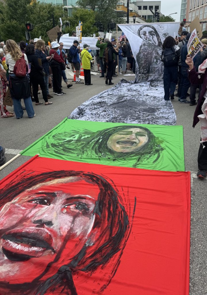Image: Kandi Jamieson's painting on fabric of a young Palestinian girl. The flags are laid down as a banner in white, green, and red backgrounds depicting faces in anguish. Image courtesy of the author.