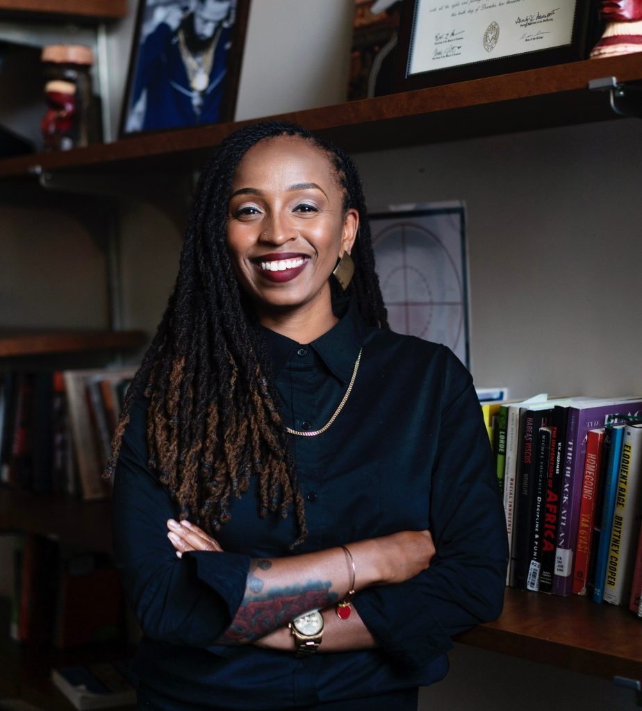 Image: A portrait of Dr. ShaDawn Battle smiling in front of a bookshelf. Courtesy of the artist.