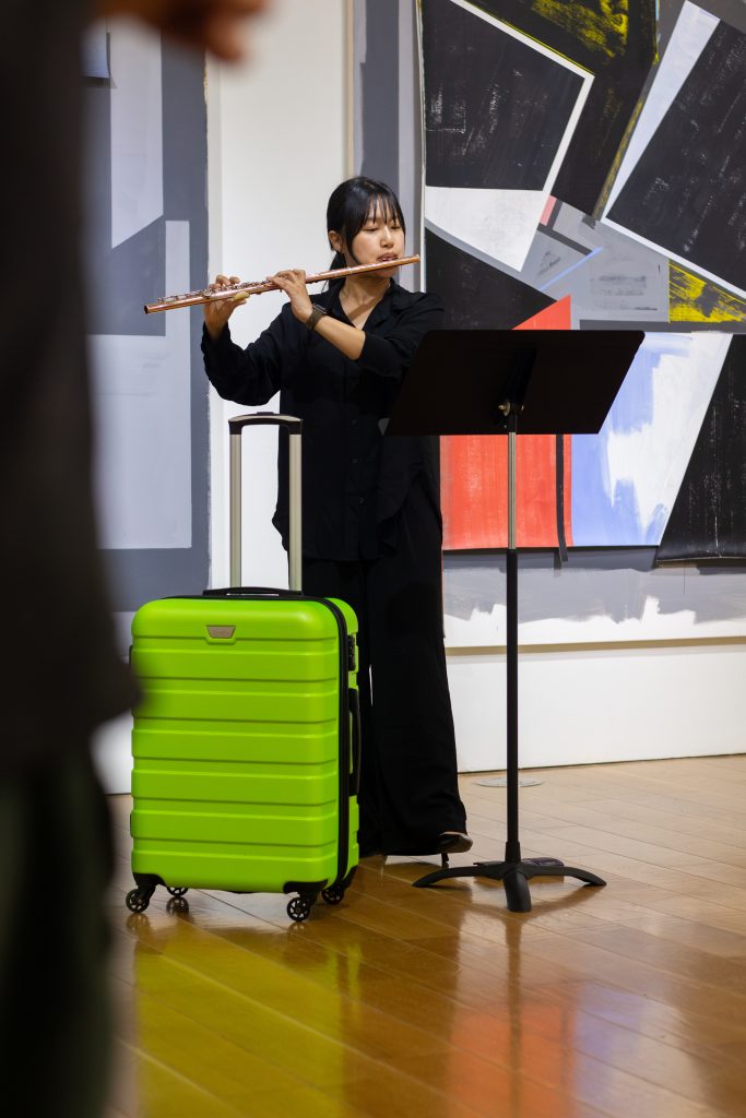 Images: (Left) Kyoungseon Lim playing a flute solo, the green suitcase at her feet, in front of Caleb Taylor’s Articulation paintings. (Right) Carlos Pacheco playing a bassoon solo behind a green suitcase, surrounded by Ariadna Ginez’s ceramic pieces and Blas Isasi’s wood and sand sculpture, 1,001,532 CE (B). Photos courtesy of Charlotte Street Foundation.