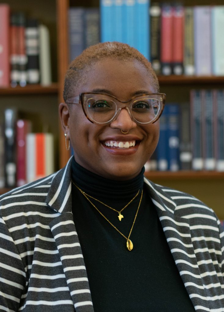 Image: Siobhan McKissic smiles at the camera while in front of a bookshelft. Image courtesy of the artist.