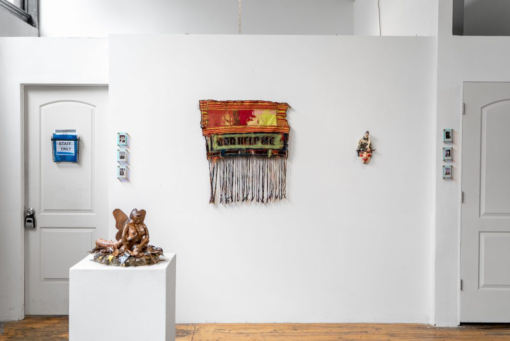 Image: Installation View, Eden Through a Glory Hole, 2025, Chicago Art Department. A sculpture on the pedestal showcases a hybrid creature or intertwining bodies and a textile in the background reads, "God Help Me." Photo: Mikey Mosher. Courtesy of Tali Halpern.