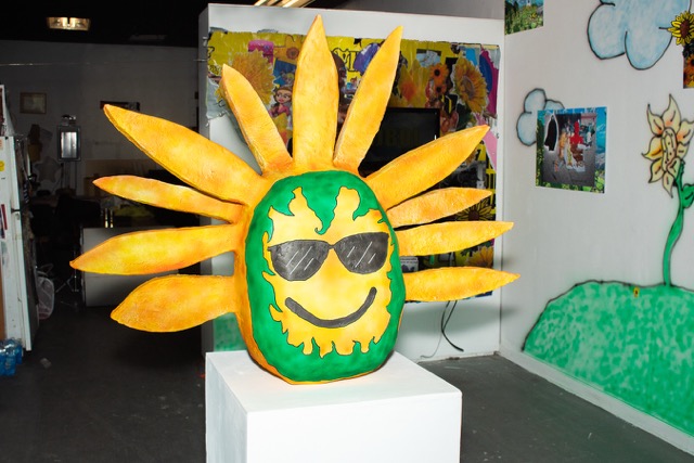 Image: Installation close up of Jujubee (sunflower/emoji idol) made of foam, paperclay, and airbrushed ink. In the foreground of the exhibit a smiling sunflower with shades and green garland around its face sits on a white pedestal. Image courtesy of the artist.
