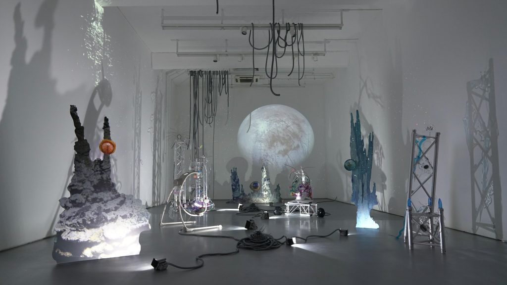 A gallery space is filled with sculptural objects that give the appearance the viewer is viewing some extraterrestrial landscape with rock formations, cables dangling from the ceiling and piled on the floor, and several architectural forms built from trusses.