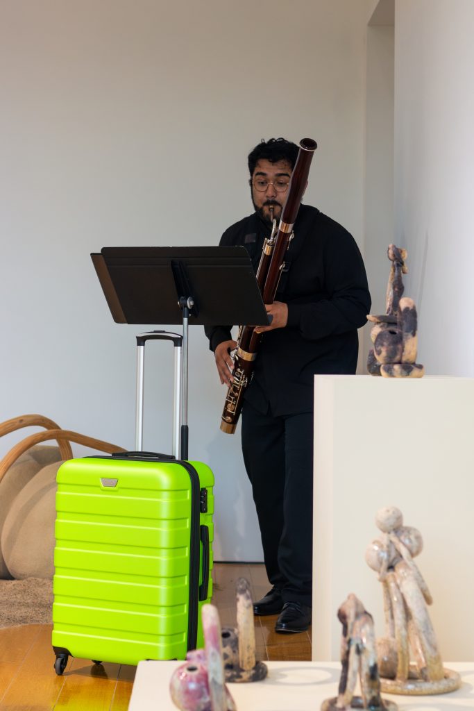 Images: (Left) Kyoungseon Lim playing a flute solo, the green suitcase at her feet, in front of Caleb Taylor’s Articulation paintings. (Right) Carlos Pacheco playing a bassoon solo behind a green suitcase, surrounded by Ariadna Ginez’s ceramic pieces and Blas Isasi’s wood and sand sculpture, 1,001,532 CE (B). Photos courtesy of Charlotte Street Foundation.