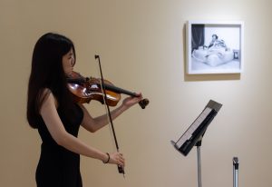 Image: Wan Loo Soo playing a viola solo while referencing her sheet music on a music stand, with Brayan Enriquez’s photograph on the wall behind. Photo courtesy of Charlotte Street Foundation.