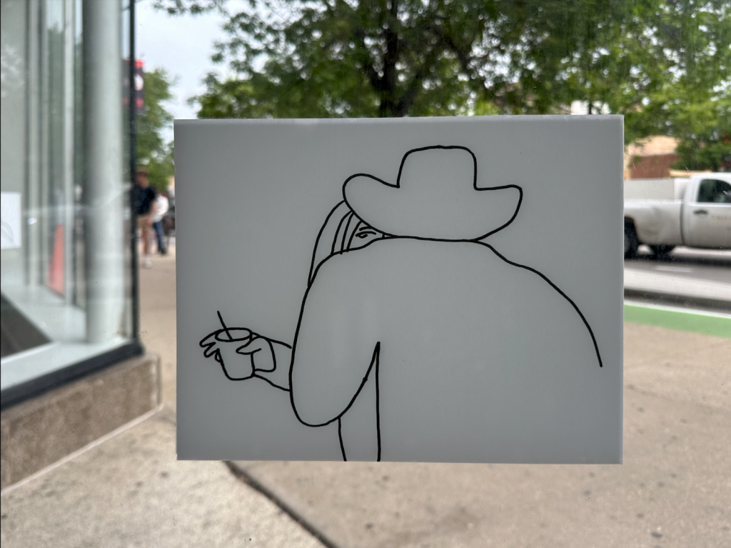 Image: (Left) A black outline drawing on paper depicts a person wearing a cowboy hat hugging or dancing with a lady holding a cup. The drawing is hung on the window glass and there’s the street scene with pedestrians in the background.