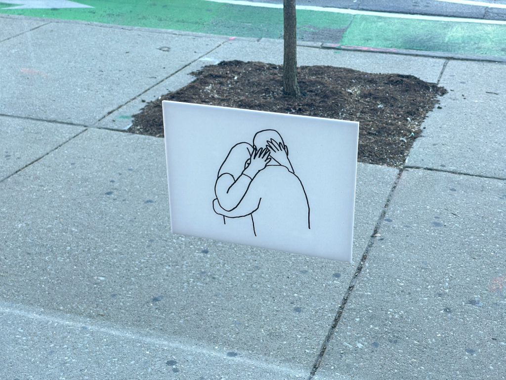 (Right) A black outline drawing on paper depicts two figures embracing or hugging with a series of simple, continuous lines. The drawing is hung on the window glass. Images courtesy of the artist.