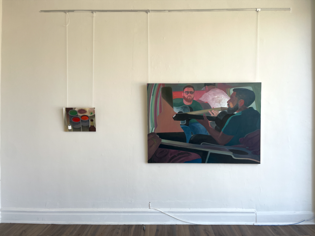 Image: A white wall is displaying two paintings. The smaller painting, hung slightly lower, shows a still life of several colored containers. The larger painting, positioned to the right, is a scene depicting multiple people sitting in a vehicle. Two figures are prominent, with one man holding or playing a guitar; the other figure is seen more distantly. The colors in the larger painting primarily consist of muted greens, purples, and browns, with darker shaded areas. The smaller painting has more vibrant tones, featuring reds, greens, and purples. Image courtesy of the artist.