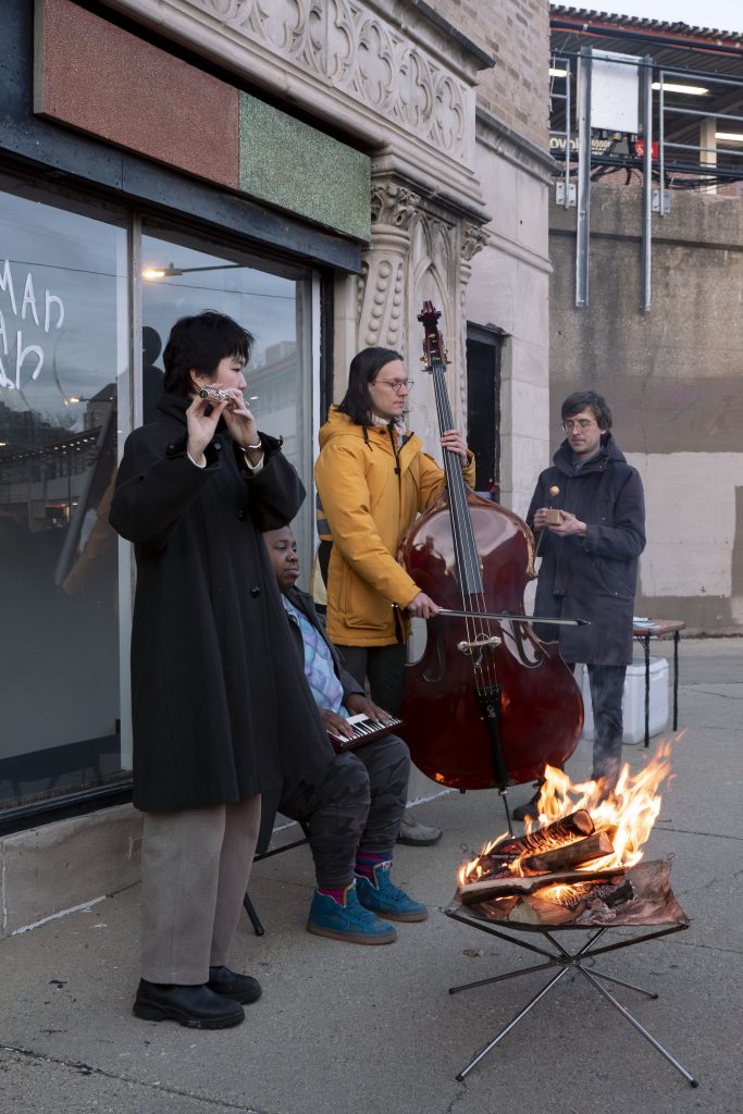 Image: Ruby Que, Eveninglessness, 2025. Four performers stand in front of a portable fire pit. They appear to be playing a variety of instruments, including a flute, keyboard, and bass. Photo by Ji Yang.
