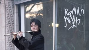 Image: Ruby Que, "Eveninglessness," 2025. A performer with cropped hair stands in front of window and holds a flute to their mouth as if playing it. The words "Roman Susan" are written in graffiti on the window. Photo by Ji Yang.