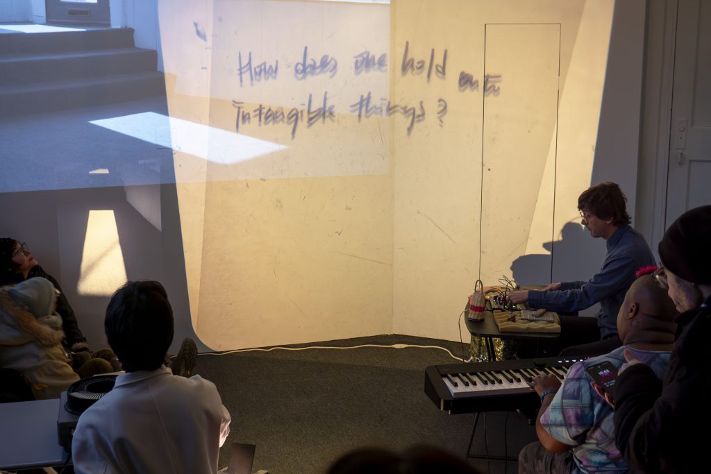 Image: Ruby Que, Eveninglessness, 2025. The phrase "How does one hold onto intangible things?" is projected onto a white gallery wall. A person operates a projector in the foreground while two musicians play synthesizers and a keyboard on the right side of the image. Audience members watch. Photo by Ji Yang.