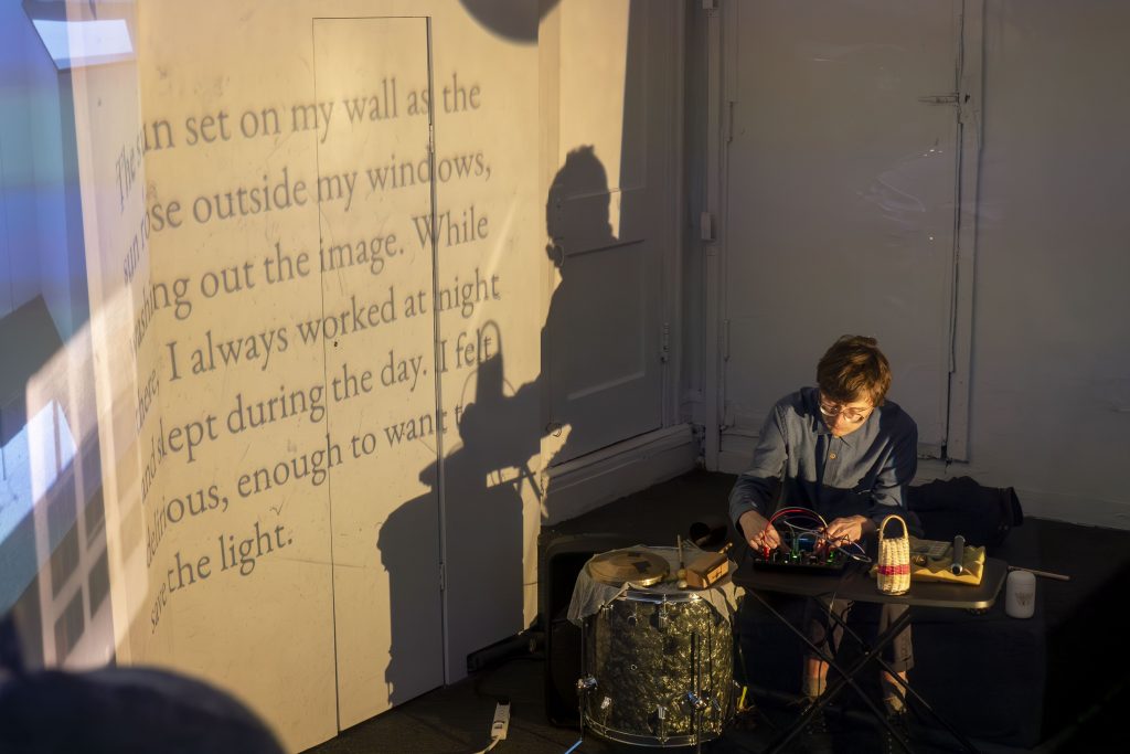 Image: Ruby Que, Eveninglessness, 2025. A performer with glasses sits at a table beside a drum and plays a synthesizer. A projection of serif text is visible on the gallery wall beside the performer. Photo by Ji Yang.
