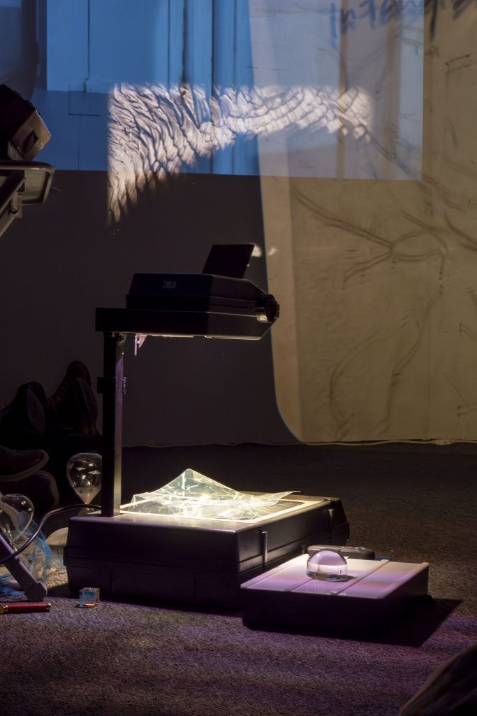 Image: Ruby Que, Eveninglessness, 2025. An overhead projector sits on the floor with a variety of objects scattered around it: a prism, hourglass, and a sheet of crinkled plastic. Indistinct and layered projections are visible in the background. Photo by Ji Yang.