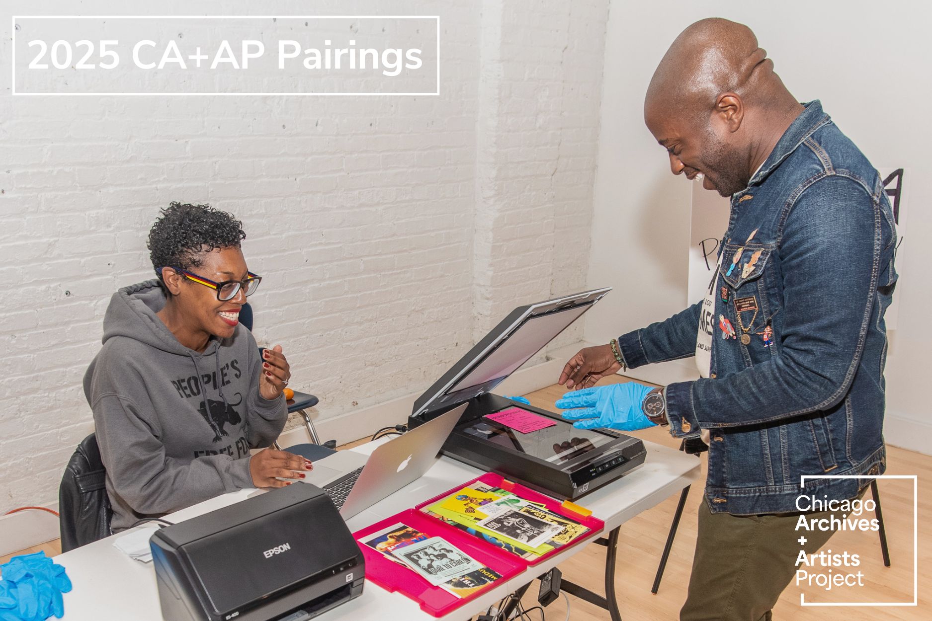 Image: Two people are at a table scanning ephemera from Honey Pot Performance's archive. Photo courtesy of Honey Pot Performance.