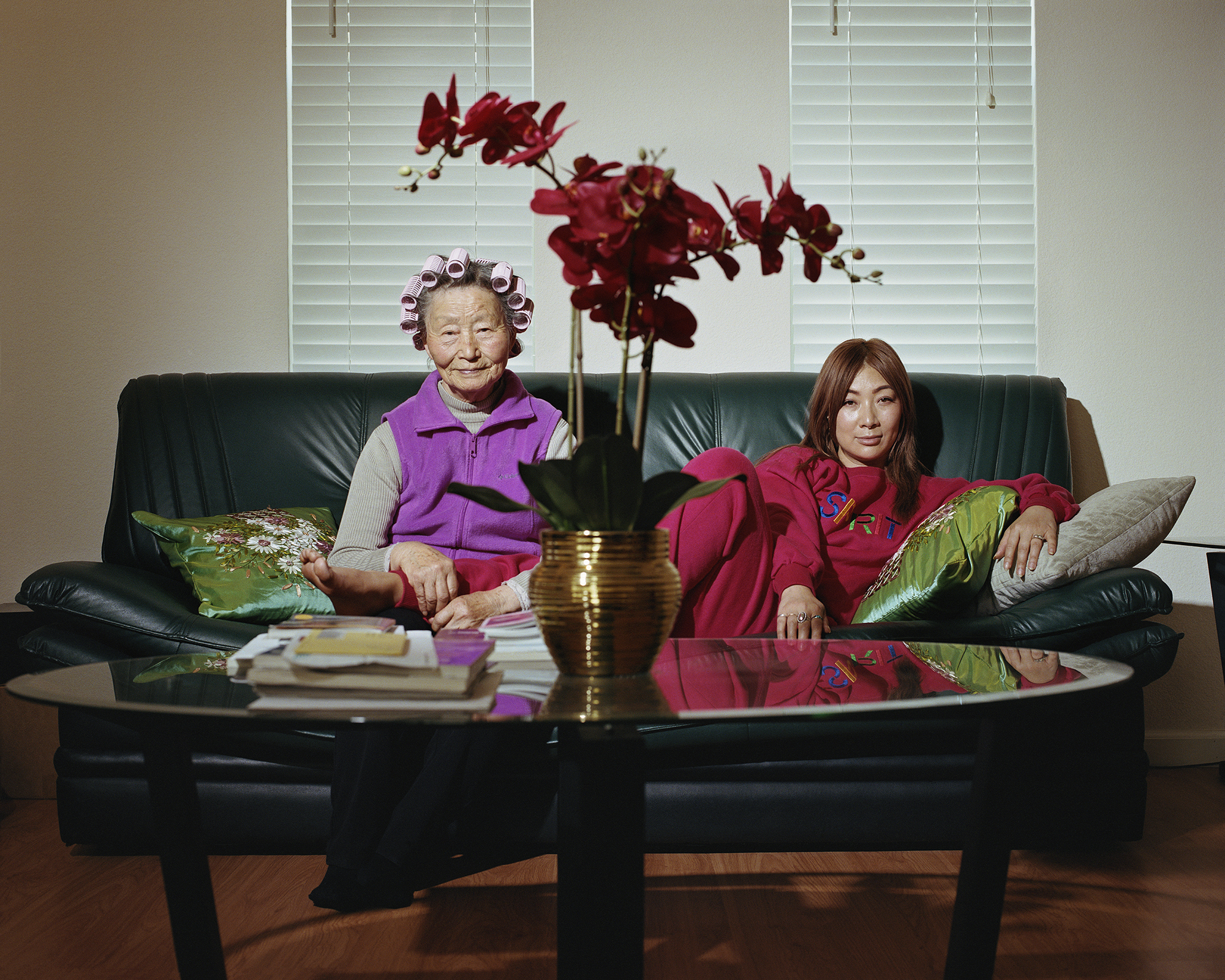 Image: Abu and Anna, 2023, Archival pigment print. Two people sit on a deep green leather couch. One is sitting upright, wears a purple vest, and has pink curlers in their hair. The other person is slouching and wears a red tracksuit. A large gold vase filled with tall red flowers sits on a coffee table in front of them. Image courtesy of the artist.
