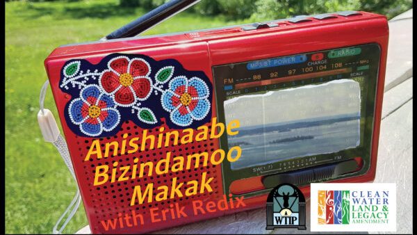 Image: A photograph of a red radio with grass in the background. Flowers are embroidered across the top left of the radio. Words are written across the top of the image that say Anishinaabe Bizindamoo Makak with Erik Redix across the front. Next to the words are logos for WTIP and Clean Water Land and Legacy Amendment on the bottom right. Image courtesy of Ojibwe Radio.
