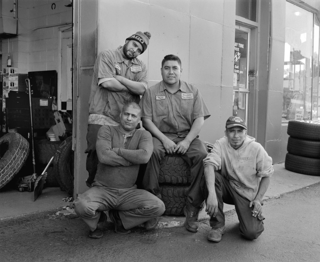 Image: “Broadway Tire Service” by Matt Schwerin. A black and white photo of four folks that presumably work at Broadway Tire Service. They pose in front of the business in various positions, with the person in the middle sitting on a stack of tires. in Photo courtesy of Belt Publishing.