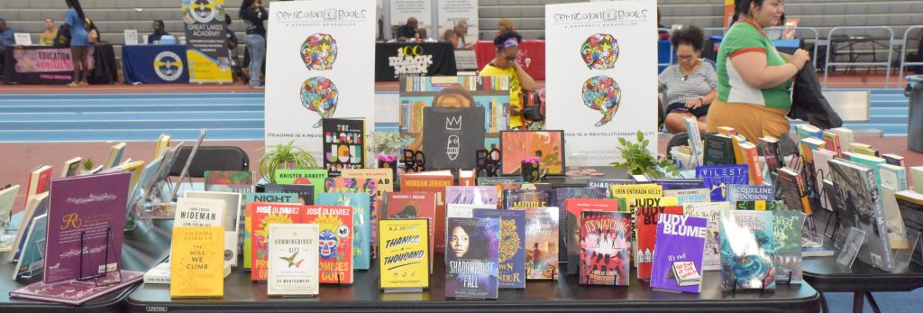 Image: In a large gym, a group of black tables arranged in a square contain rows of books with colorful covers. In the background, people chat near tables and read books. Photo created by EdVetté Wilson Jones.