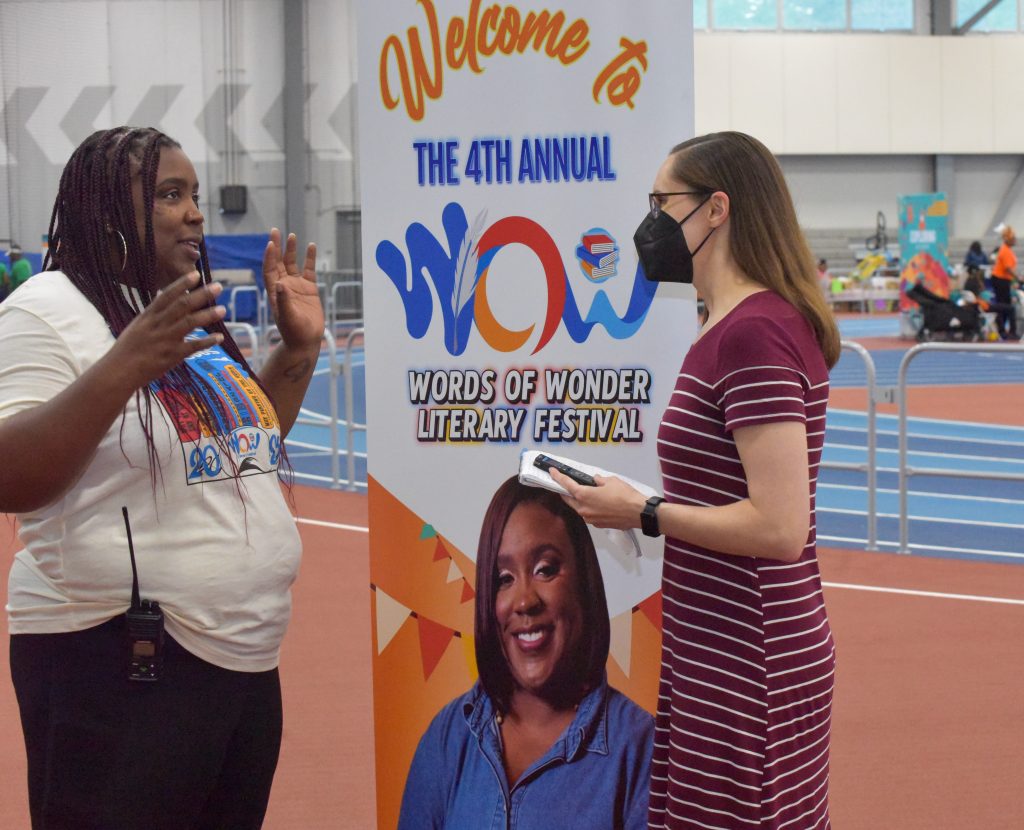Image: Burst Into Books founder Jurema Gorham and Emily McClanathan chat in front of a poster board that reads "Welcome to The 4th Annual Words of Wonder Literary Festival" inside of a gymnasium. Photo by Edvétte Wilson Jones.