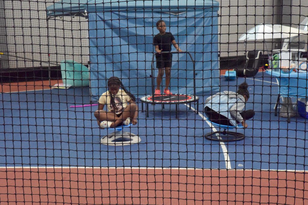 Image: Three children bounce and play on trampolines in a school gym, seen through wall of black netting. Photo by EdVetté Wilson Jones.
