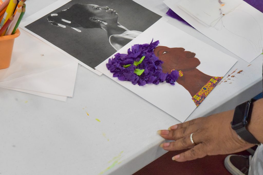 Image: On a white table with colored pencils, a black and white image of blues singer Nina Simone with a painted replica of the image underneath. A brown hand grazes the edge of the table. Photo by EdVetté Wilson Jones.