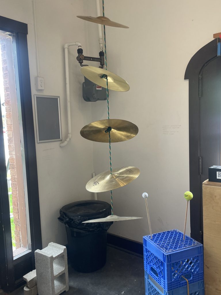 Image: In the corner of Comfort Station near the entrance, cymbals hang on a rope vertically from the ceiling in spaced out tiers. Sticks—one with a golf ball ball at the top, one with a tennis ball at the top—are stuck in a crate to hit the cymbals with. Image by Nadia John.