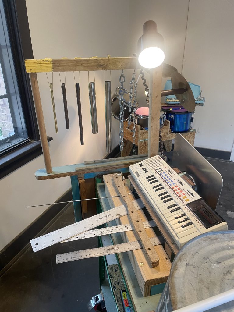 Image: A cart full of moveable instruments including chimes, rulers to pluck, a trash can drum, and an electric keyboard. Behind this cart a homemade drum set can be seen. The cart is illuminated by a spotlight lamp attached to the chimes. Image by Nadia John.